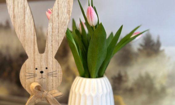 Ein Holzhase steht neben einer Vase mit Tulpen auf einem Tisch. Der Frühling ist da. Die Sonne scheint durch das Fenster.