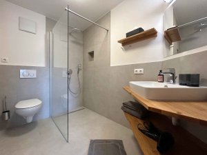 In einem Badezimmer stehen eine Dusche, ein Waschbecken und eine Toilette. Es gibt Regale und einen Handtuchhalter.