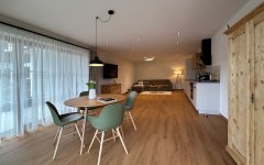 Ein Raum zeigt einen Essbereich mit Tisch, Stühlen und einer Küche. Es gibt Fenster mit Vorhängen und Holzfußboden.