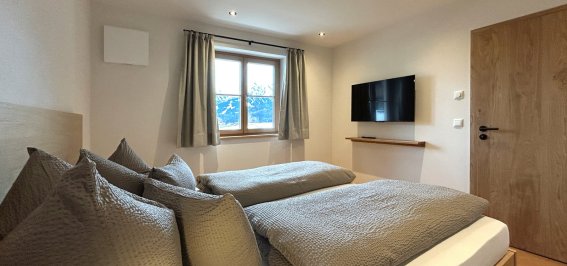 Gemütliches Zimmer mit zwei Betten in den Bergen Das Zimmer zeigt zwei Einzelbetten, einen Fernseher und einen Blick auf die Berge. Tageslicht strömt durch das Fenster.