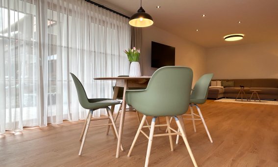 Ein Essbereich zeigt einen Tisch mit Stühlen. Tageslicht strömt durch die Fenster. Ein Wohnzimmer ist im Hintergrund sichtbar.