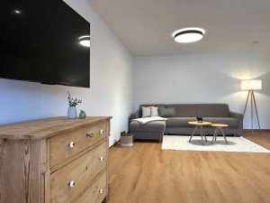 In einem Wohnraum steht ein graues Sofa vor einem großen Fernseher. Ein Tisch und eine Lampe sind auch im Raum.