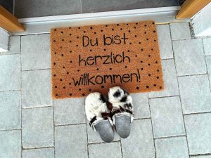 Die Fußmatte sagt Willkommen. Daneben liegen Hausschuhe auf dem Boden. Es ist ein klarer Tag.