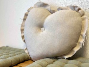 Ein Kissen in Herzform liegt auf einem Tisch. Es ist aus einem grauen Stoff und hat eine Druckknopfmitte.
