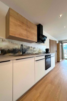 Die Küche zeigt Holzregale und moderne Geräte. Natürliches Licht kommt von einem Fenster. Der Raum ist schlicht und funktional.