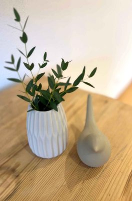 Auf einem Tisch stehen eine Vase mit grünen Zweigen und eine Vogelfigur aus Keramik. Die Szene ist einfach und klar.
