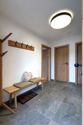 Im Flur stehen eine Sitzbank und ein Garderobenständer. Es gibt Holz- und Fliesenbelag. Die Türen sind aus Holz.