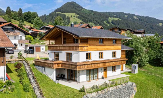 Ein modernes Haus steht in den Bergen. Es gibt viele Bäume und andere Gebäude in der Nähe. Der Himmel ist blau und die Landschaft ist grün.