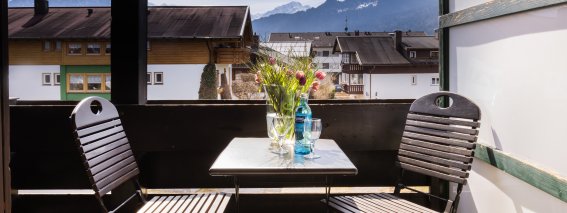 Auf dem Balkon stehen zwei Stühle und ein Tisch. Im Hintergrund sieht man Berge und einen klaren Himmel.