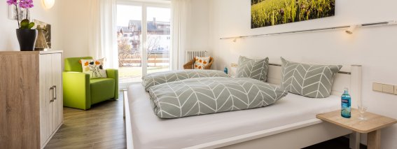 Das Zimmer zeigt ein bequemes Bett und eine grüne Sitzecke. Es gibt eine große Fensterfront mit Blick nach draußen.