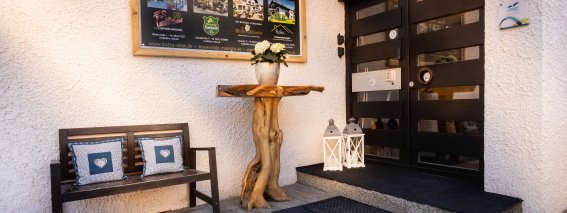 Der Eingang zeigt eine Holzbank und einen Tisch mit Blumen. Es gibt eine große Tafel mit Informationen und das Wetter ist schön.