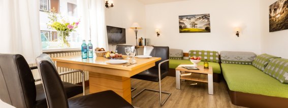 In einem Wohnzimmer stehen ein Tisch mit Snacks und Getränke. Die Sitzecke hat eine Couch und schwarze Stühle.