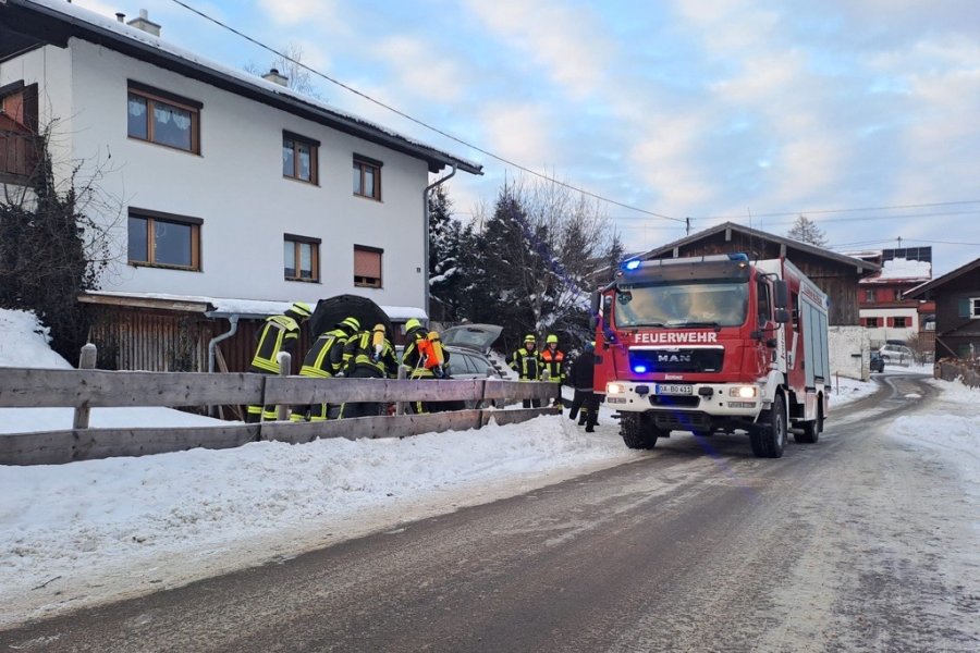 B2 - Brand PKW Feuerwehrleute kontrollieren einen PKW nach einem Brand im Motorraum in einer verschneiten Straße. Ein Feuerwehrlöschfahrzeug steht in der Nähe.