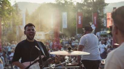 Ein Musiker spielt Gitarre auf einer Bühne vor vielen Menschen bei einem Event in der Abendsonne im Freien.