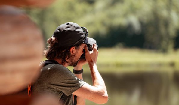 Ein Mann macht Fotos an einem See Ein Mann steht am Ufer eines Sees und richtet die Kamera auf ein Motiv. Es ist tagsüber und die Natur ist sichtbar.