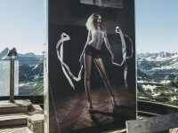 Ein großes Ausstellungsbild steht auf der Nebelhorn Terasse. Auf dem Bild ist eine Frau mit fliegenden Bändern in der Hand zu sehen. Im Hintergund hängt ein Basketballkorb. Hinter derm Ausstellungsbild ist das Bergpanorama der Allgäuer Alpen zu sehen.