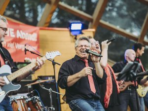 Eine Band spielt Live-Musik im Kurpark in der Muschel. Der Auftritt findet am Abend statt und es gibt mehrere Musiker. Die Stimmung ist gut.