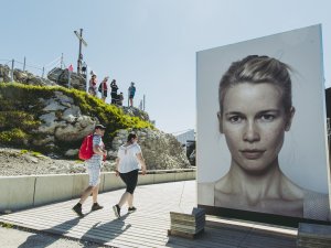 Unterhalb des Nebelhorner Gipfelkreuzes steht ein großer Aufsteller mit einem Bild der Fotogipfel Austellung. Dort ist ein Portrait einer Frau zu sehen.