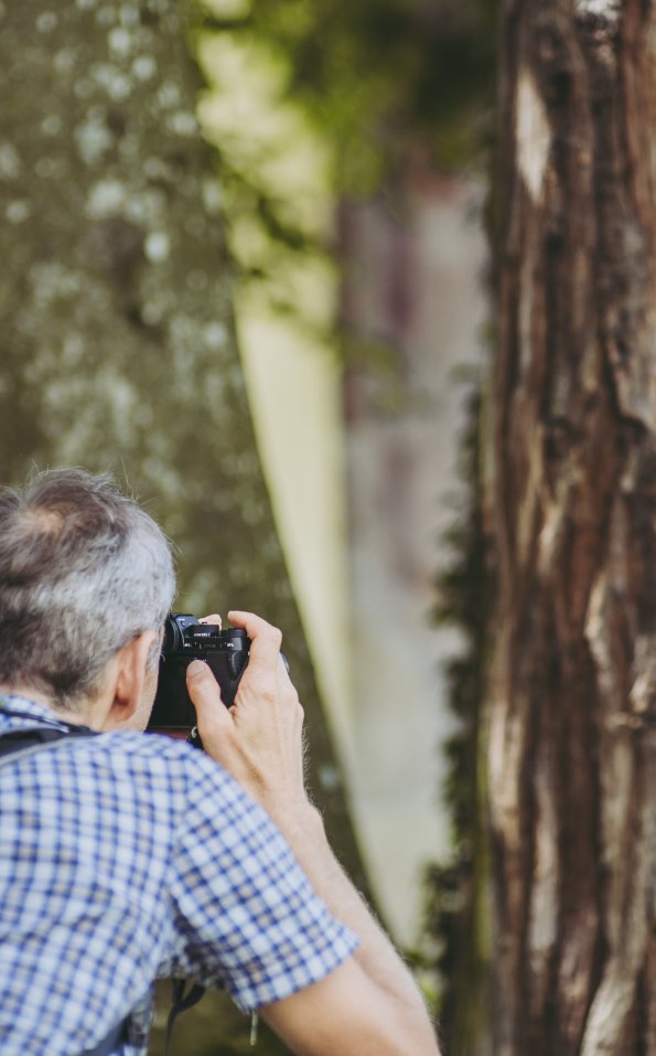 Ein Teilnehmer des Fotogipfel Fotographie-Workshops steht vor einem Baum und hält eine Kamera. Er macht Aufnahmen in der Natur.