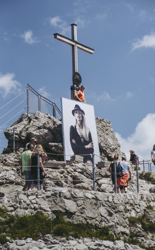 Direkt am Gipfelkreuz des Nebelhorns steht ein Ausstellungsbild des Fotogipfels. in dieser spektakulären Location wird eine Frau mit schwarzer Kleidung und Eindrucksvoller Haltung gezeigt.