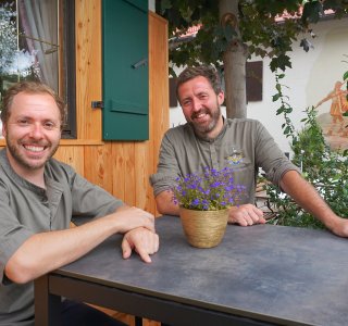 Zwei Köche sitzen an einem Tisch im Freien und lächeln. Sie befinden sich in einer ruhigen Umgebung mit Pflanzen.