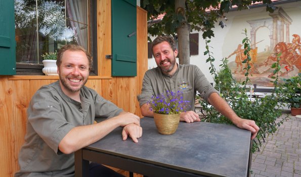 Zwei Köche sitzen an einem Tisch im Freien und lächeln. Sie befinden sich in einer ruhigen Umgebung mit Pflanzen.