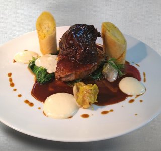 Das Gericht zeigt Fleisch mit Soße, Gemüse und Beilagen auf einem weißen Teller. Es ist ein aufwendiges Essen.