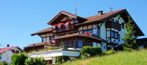 Das Haus steht in einem Tal. Der Himmel ist blau. Der Rasen ist grün. Blumen sind auf dem Balkon.