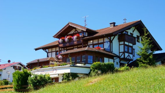 Das Haus steht in einem Tal. Der Himmel ist blau. Der Rasen ist grün. Blumen sind auf dem Balkon.