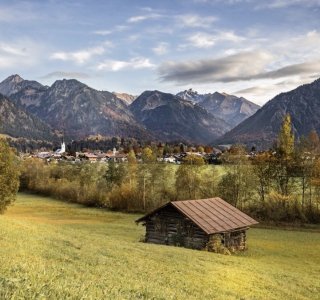 Ein Haus in einer Hügellandschaft in den Alpen Ein Holzhaus steht in einer Wiese mit Blick auf die Alpen. Die Landschaft zeigt Bäume, Berge und einen kleinen Ort.