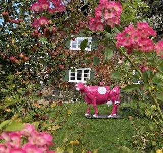 Ein Garten mit vielen Blumen und einer rosa Kuh steht vor einem alten Haus. Es ist ein sonniger Tag und alles blüht.