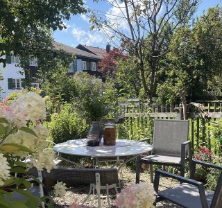 Ein Tisch mit Stühlen steht im Garten. Die Sonne scheint. Blumen und Bäume sind im Hintergrund sichtbar.