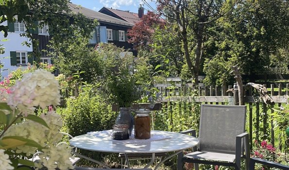 Gartenplatz unter blauem Himmel mit Blumen Ein Tisch mit Stühlen steht im Garten. Die Sonne scheint. Blumen und Bäume sind im Hintergrund sichtbar.