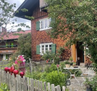 Ein Haus mit Holzfassade steht in einem Garten. Blumen blühen und ein Tisch steht auf der Terrasse.