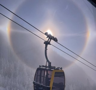 Seilbahn fährt durch einen Lichtkreis in den Bergen Eine Seilbahn fährt auf einen Berg. Ein Lichtkreis um die Sonne ist zu sehen. Der Tag ist klar und kühl.