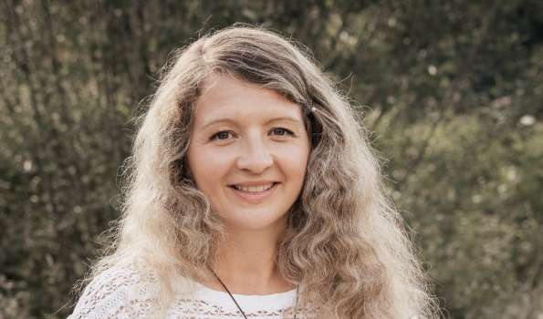 Silke Eine Frau mit lockigem Haar steht im Freien zwischen Pflanzen und schaut in die Kamera. Es ist tagsüber und sonnig.