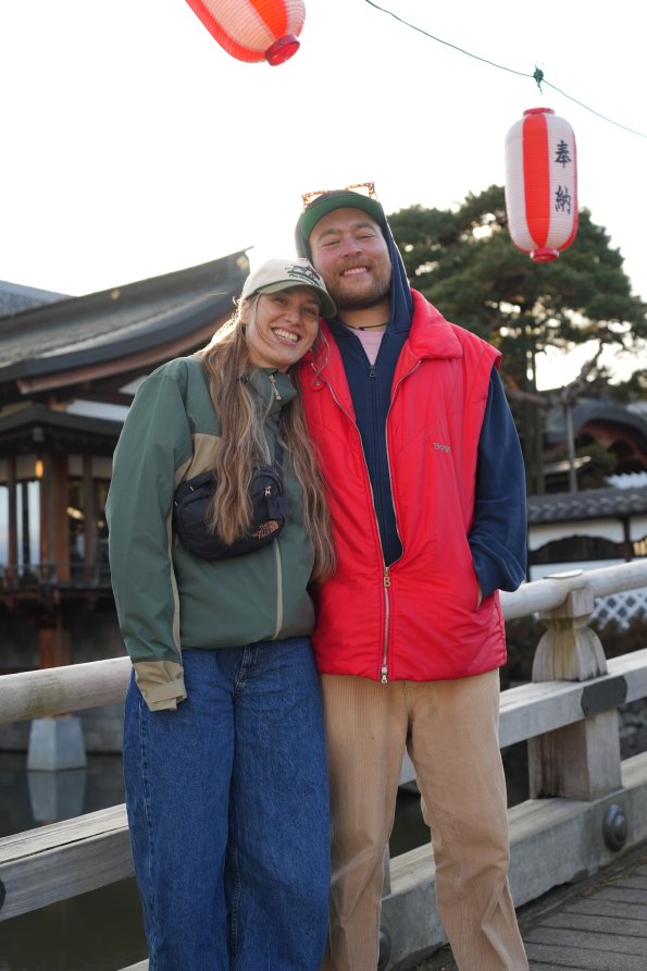 Marzellus und Antonia stehen auf einer Brücke in Japan. Sie lächeln und tragen bequeme Kleidung. Es ist Abend und Licht leuchtet.
