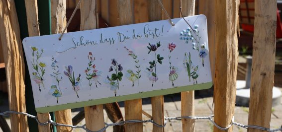 Das Schild begrüßt Besucher mit einem freundlichen Spruch und zeigt verschiedene Pflanzenmuster in einem Gartenbereich.