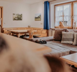 Im Raum gibt es eine Couch, einen Tisch und Bilder an der Wand. Ein Hund sitzt im Vordergrund und schaut zur Kamera.