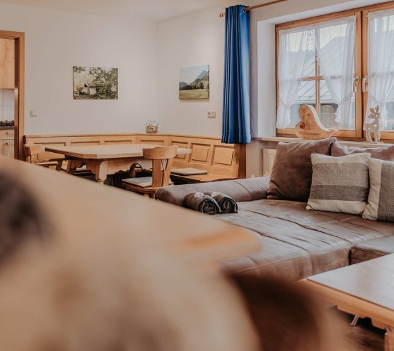 Im Raum gibt es eine Couch, einen Tisch und Bilder an der Wand. Ein Hund sitzt im Vordergrund und schaut zur Kamera.