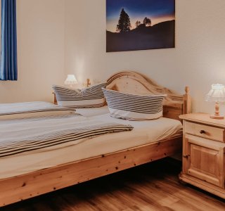 In einem Zimmer ein Doppelbett. Es gibt einen Nachttisch und ein Fenster, das Licht lässt herein.