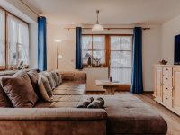 In einem Raum steht ein großes Sofa. Vor den Fenstern hängen blaue Vorhänge. Ein Tisch steht in der Mitte.