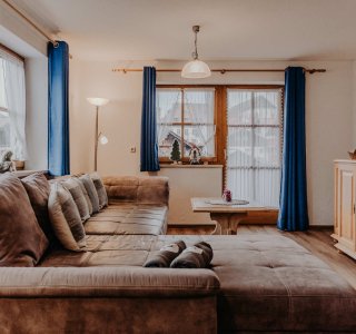 In einem Raum steht ein großes Sofa. Vor den Fenstern hängen blaue Vorhänge. Ein Tisch steht in der Mitte.