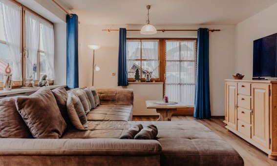 In einem Raum steht ein großes Sofa. Vor den Fenstern hängen blaue Vorhänge. Ein Tisch steht in der Mitte.