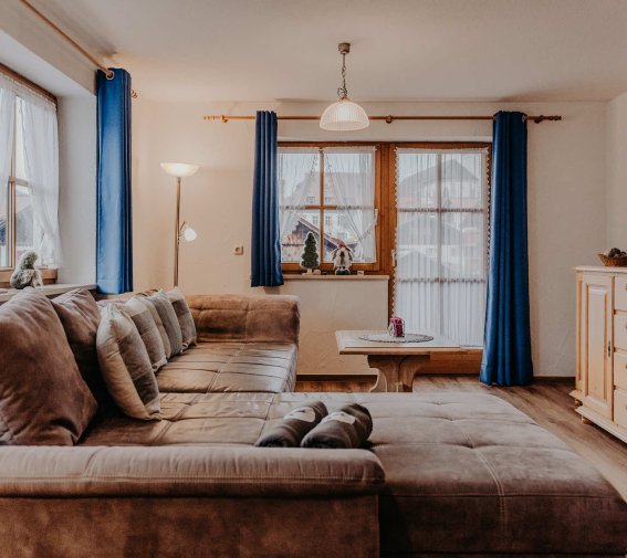 Wohnzimmer mit einem großen Sofa und Fenster In einem Raum steht ein großes Sofa. Vor den Fenstern hängen blaue Vorhänge. Ein Tisch steht in der Mitte.