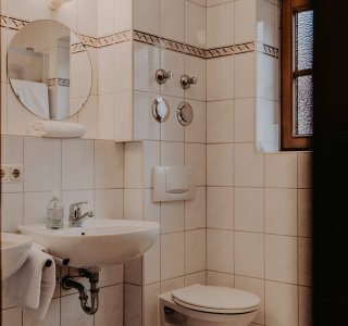 In einem Badezimmer stehen ein Waschbecken und eine Toilette. Es gibt einen Spiegel und eine kleine Fensteröffnung.