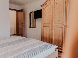 In einem Raum gibt es ein Bett, einen Holzschrank und einen Spiegel an der Wand. Der Raum wirkt gemütlich und klar.