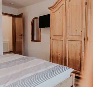 In einem Raum gibt es ein Bett, einen Holzschrank und einen Spiegel an der Wand. Der Raum wirkt gemütlich und klar.