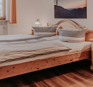 Das Schlafzimmer hat ein großes Bett mit Kissen. Es gibt einen Nachttisch und ein Fenster. Es ist Tag und das Licht scheint.