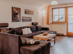 Ein Wohnzimmer mit einem großen Sofa und einem Tisch. Die Fenster lassen viel Licht herein. Es ist eine ruhige Atmosphäre.